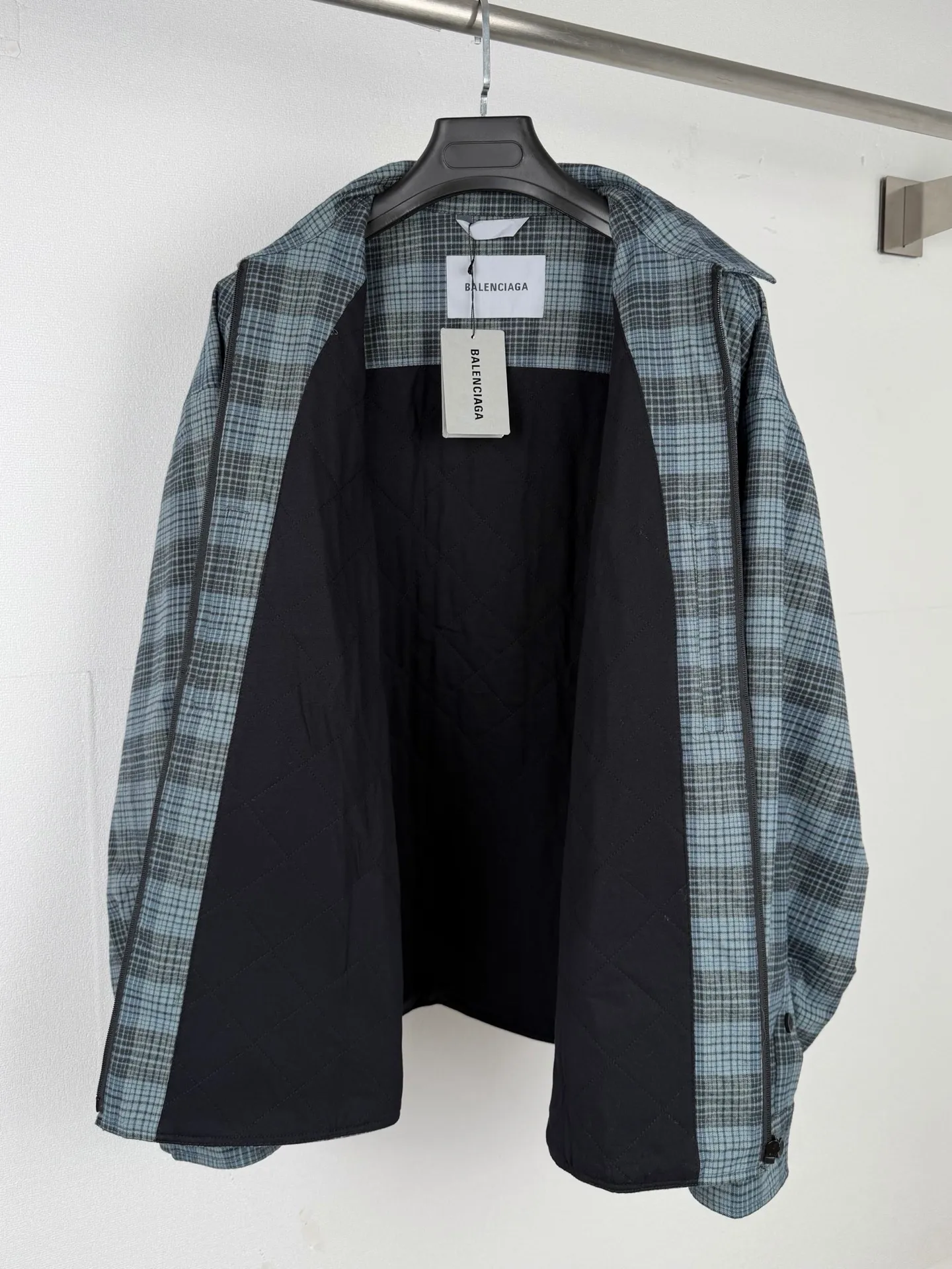 BALENCIAGA/balenciaga blue and gray plaid zipper pocket logo quilted jacket combines a three-layer structure of plaid woven fabric, filling cotton, and lining, and integrates plaid alignment technology and quilted garment production standards. There are multiple process difficulties in the whole process. The surface layer of the body is made of blue-gray Retro plaid fabric, and the cutting machine production strictly follows the global grid standards. The arrangement direction of the cutting pieces is fixed and cannot be changed at will. The horizontal and vertical lines of shoulder seams, side seams, and zipper placket joints must be consistent and coherent. The left chest patch pocket is cut independently, and the bag check pattern must be aligned with the front reference pattern. Slight deviations will destroy the visual integrity and directly increase fabric loss and cutting time. The most difficult part of the style is the three-layer composite sewing. The outer plaid fabric, filling cotton, and lining fabric have different physical properties, and the preshrinking processing parameters need to be adjusted separately. Pressure and temperature control are crucial during lamination operations. Once the parameters are out of balance, it can easily cause fabric stretch and distortion, plaid dislocation, and wrinkles and bulges in garments. At the same time, it is necessary to reasonably plan the quilting line to avoid the visual beauty of the grid pattern from being cut by the lines on the basis of firmly fixing the cotton layer and preventing cotton from running away. The placket adopts a centered zipper structure. It is difficult to sew the zipper tape and the quilted panels. It is required that the width of the left and right plackets be equal, the zipper routing is straight and smooth, and there is no lag in opening and closing. When the shirt lapel is filled with cotton, the thickness of the cotton needs to be controlled to prevent the collar from becoming bloated and unnaturally folded. The metal nameplate of the chest pocket is sewn at fixed points to avoid pulling the fabric during the operation and ensure that the plaid pattern is smooth and not deformed. The quilted material increases the overall thickness of the fabric. When sewing, you need to continuously adjust the presser foot pressure and stitch length to adapt to changes in sandwich thickness. The low-temperature and gentle ironing process is used for post-finishing to prevent the plaid from fading and the cotton layer from clumping due to high temperatures. From the precise alignment of cutting pieces, three-layer fabric pretreatment, composite quilting to garment finalization inspection, the processes are interlocking and the error tolerance rate is extremely low, testing the accuracy of plate making, equipment control and turning technology. Size:XS S M L